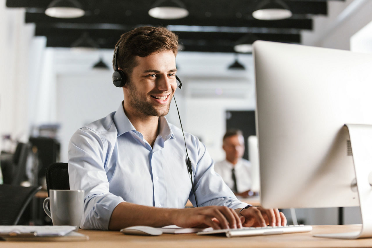 Gut gelaunter junger Mann sitzt mit Headset an seinem Schreibtisch