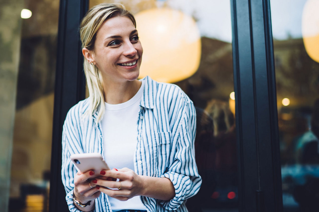 Junge, gut gelaunte Frau steht lächelnd vor einem Café und hält ihr Smartphone in der Hand