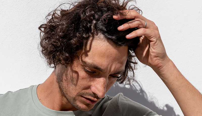 A man standing against a white wall in the sun running his hand through his curly brown hair while looking down